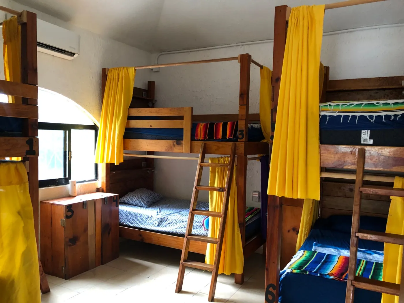 A bright hostel dorm with wooden bunk beds, yellow privacy curtains and colourful blankets
